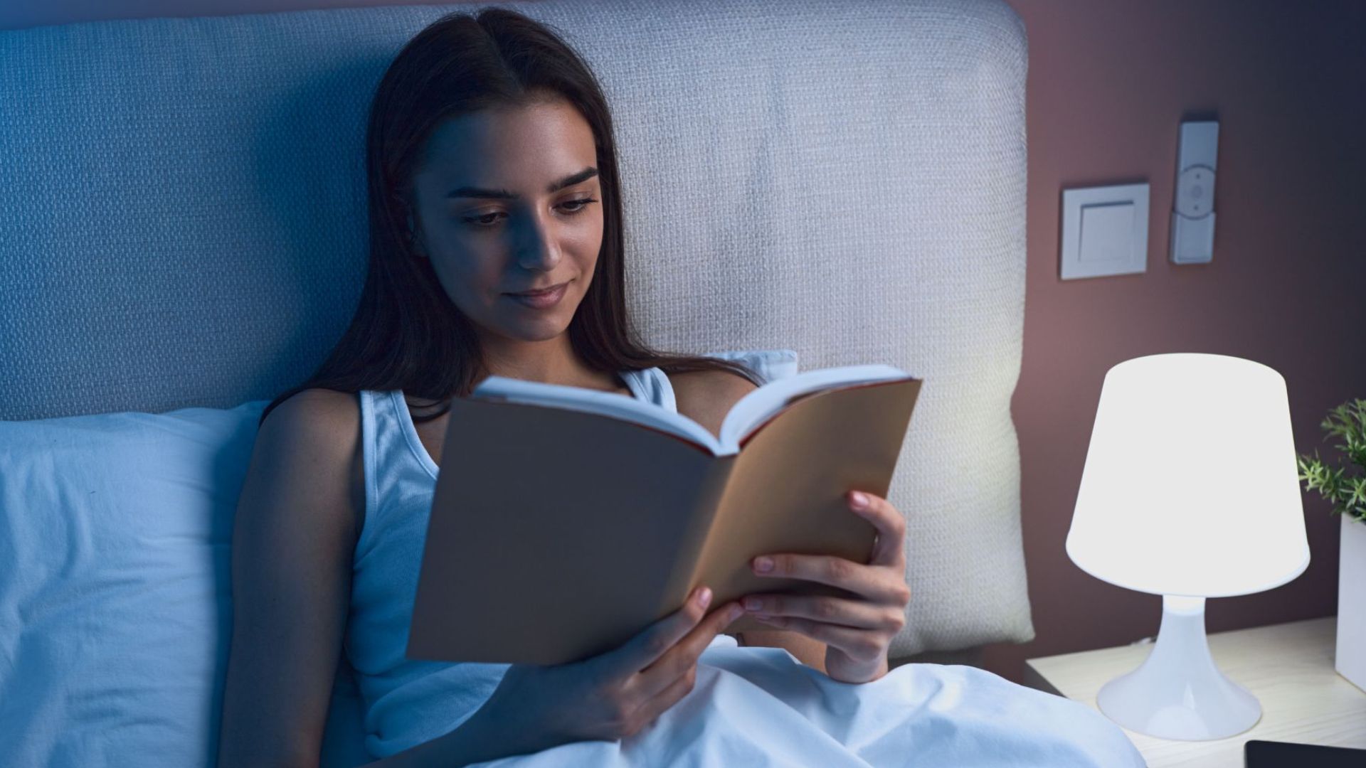 Woman sits up in bed reading with a light on her bed stand next to her.