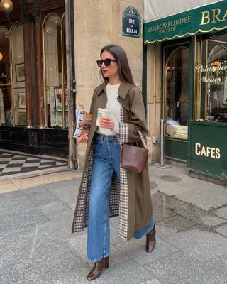 Influencer wears a trench coat with a checkered lining, a brown bucket bag, ivory sweater, blue jeans, and brown boots.