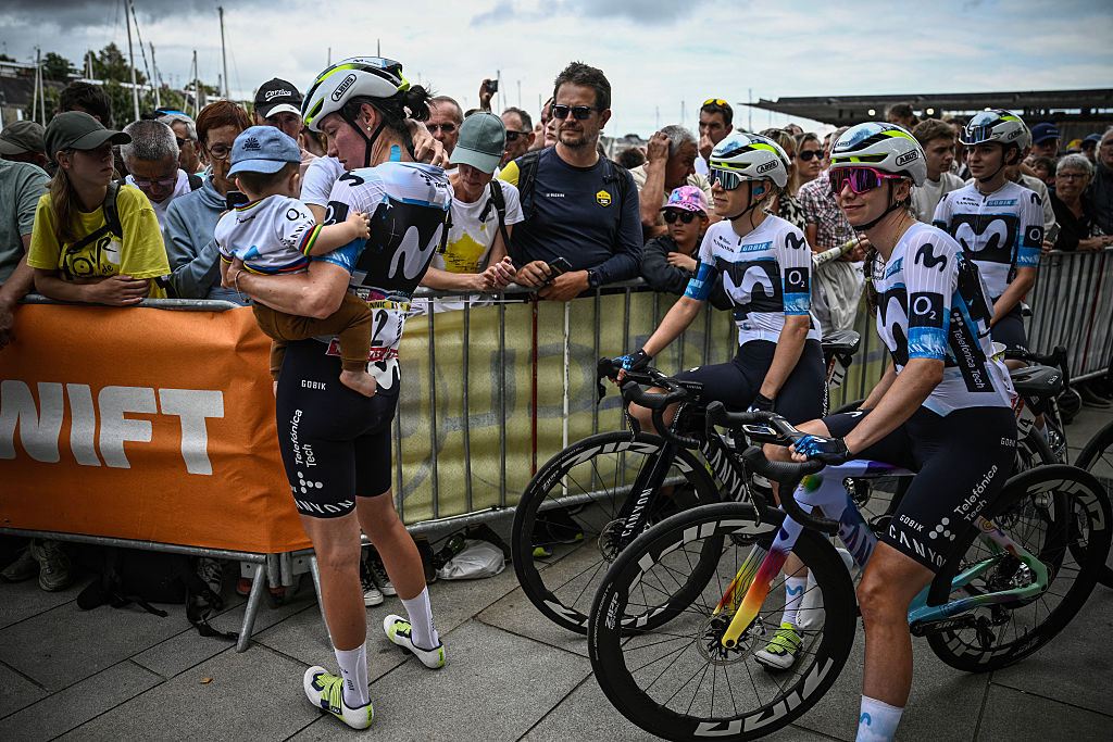 Movistar Team's French rider Aude Biannic (L) holding her son Noah in her arms waits for the presentation of the teams prior to the 1st stage (out of 9) of the fourth edition of the Women's Tour de France cycling race, 78,8 km from Vannes to Plumelec, in Vannes, western France, on July 26, 2025. (Photo by JULIEN DE ROSA / AFP)