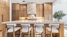 Natural wood cabinetry in a wood and warm marble kitchen, with floor to ceiling cabinetry and large island with four white upholstered bar stools. There is a potted tree visible in the background