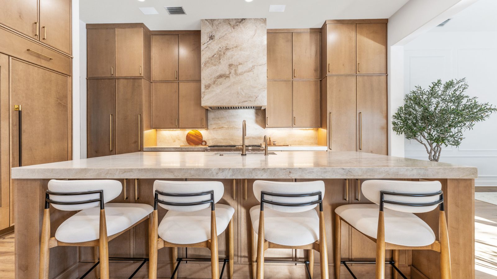 Natural wood cabinetry in a wood and warm marble kitchen, with floor to ceiling cabinetry and large island with four white upholstered bar stools. There is a potted tree visible in the background
