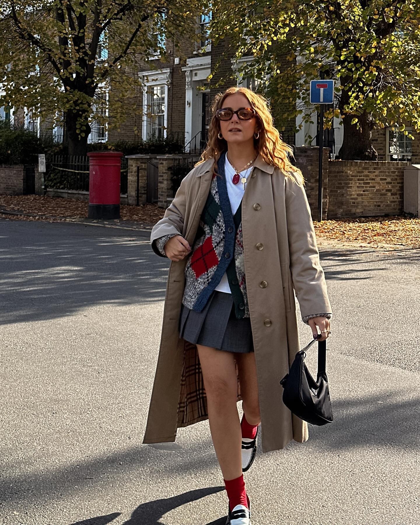 Influencer @rebeccaferrazwyatt walks down the street outside wearing an argyle cardigan over a white tee with a pleated miniskirt and beige trench coat.