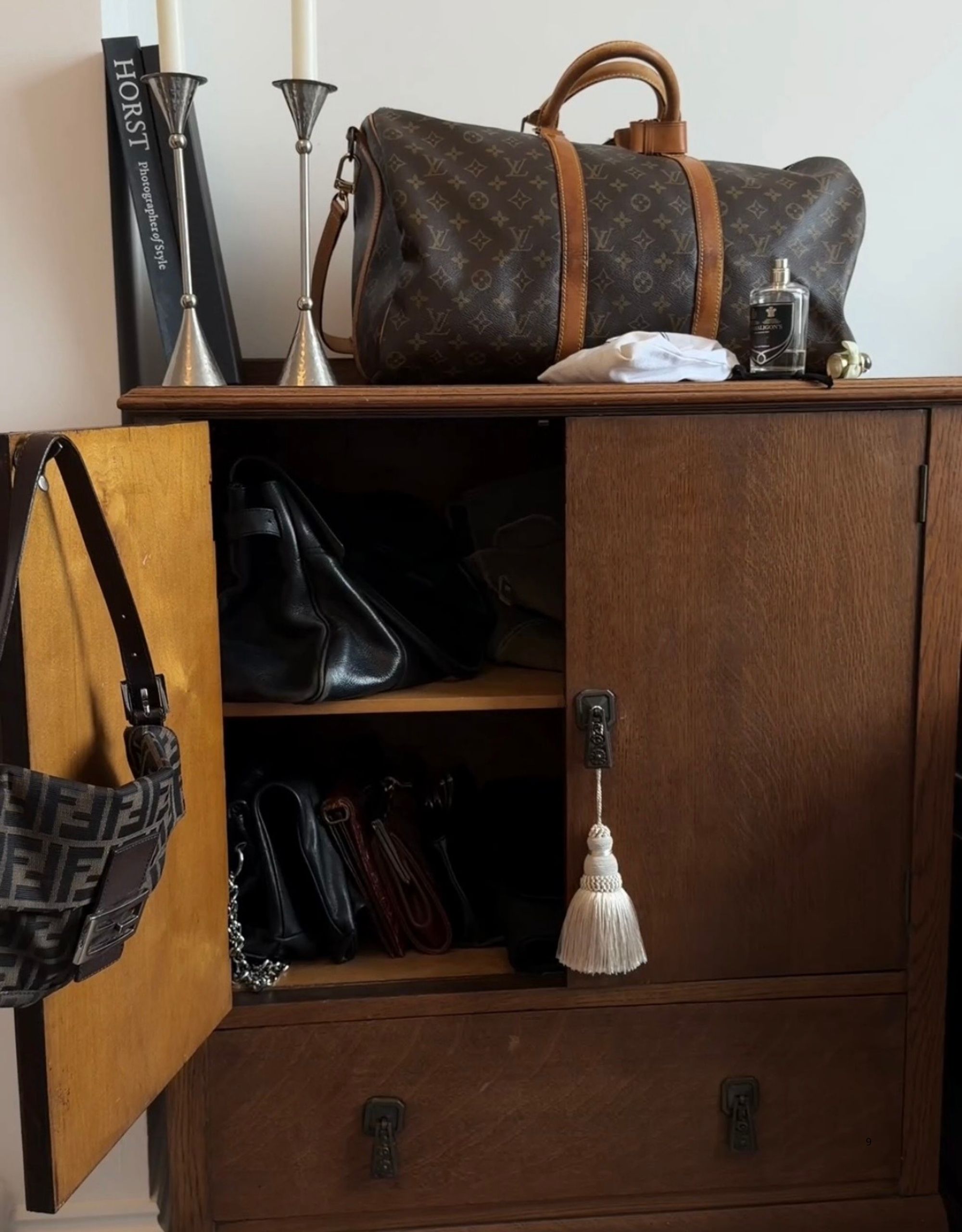 Image of a wooden cabinet open showing it's filled with purses. There is a purse hanging on the open door and a luggage bag, along with a pair of silver candlestick holders, on top of the cabinet.