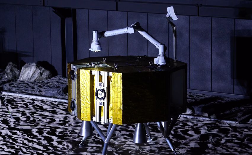 a gold-foil-wrapped octagonal lunar probe is landed on a grey surface. A white robotic arm is attached to the probe&#039;s flat top, next to an antenna.