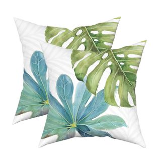 Two white square outdoor throw pillows with a green monstera leaf on the top right corner and a turquoise blue flower on the left hand corner