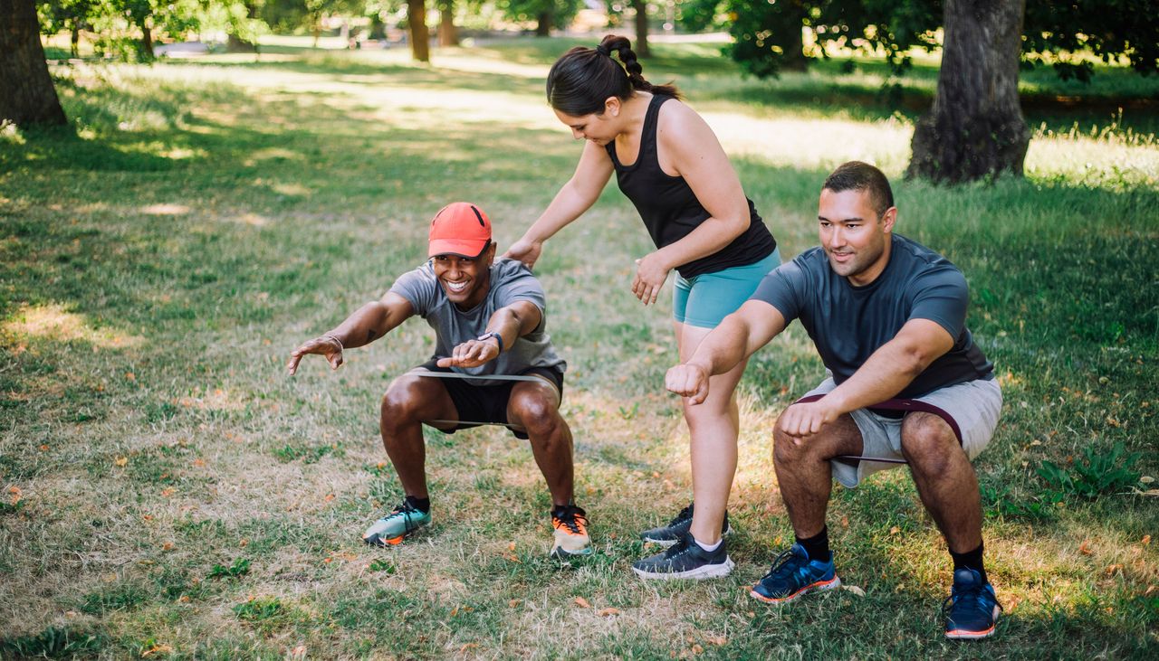 Two men squat in a park while a personal trainer corrects one man&#039;s form