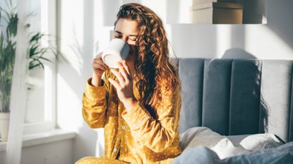 Woman in yellow pajama sitting on bed and drinking coffee at the sunny morning bedroom. - stock photo