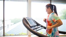 woman wearing a tie-dyed tshirt on a treadmill in front of large windows, sideways to the camera.