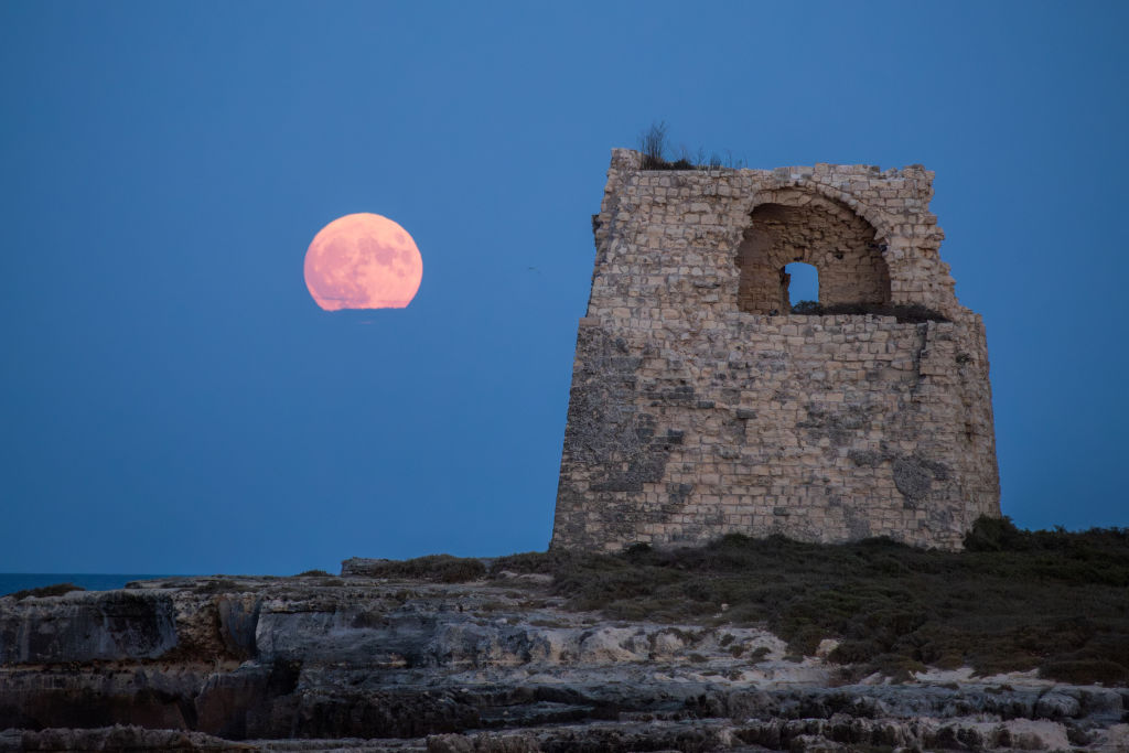 Bulan biru raksasa di sebelah kiri bersinar merah muda di samping struktur menara batu dengan bukaan kecil di bagian atasnya.