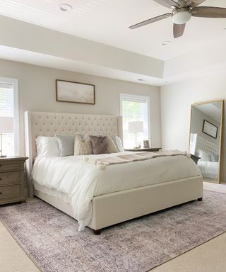 White master bedroom with button-back upholstered headboard, mirror leaning against wall, and large area rug in subtle pattern.