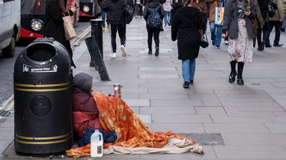 Shoppers and visitors on Oxford Street walk past a homeless man sitting against a rubbish bin while asking for money