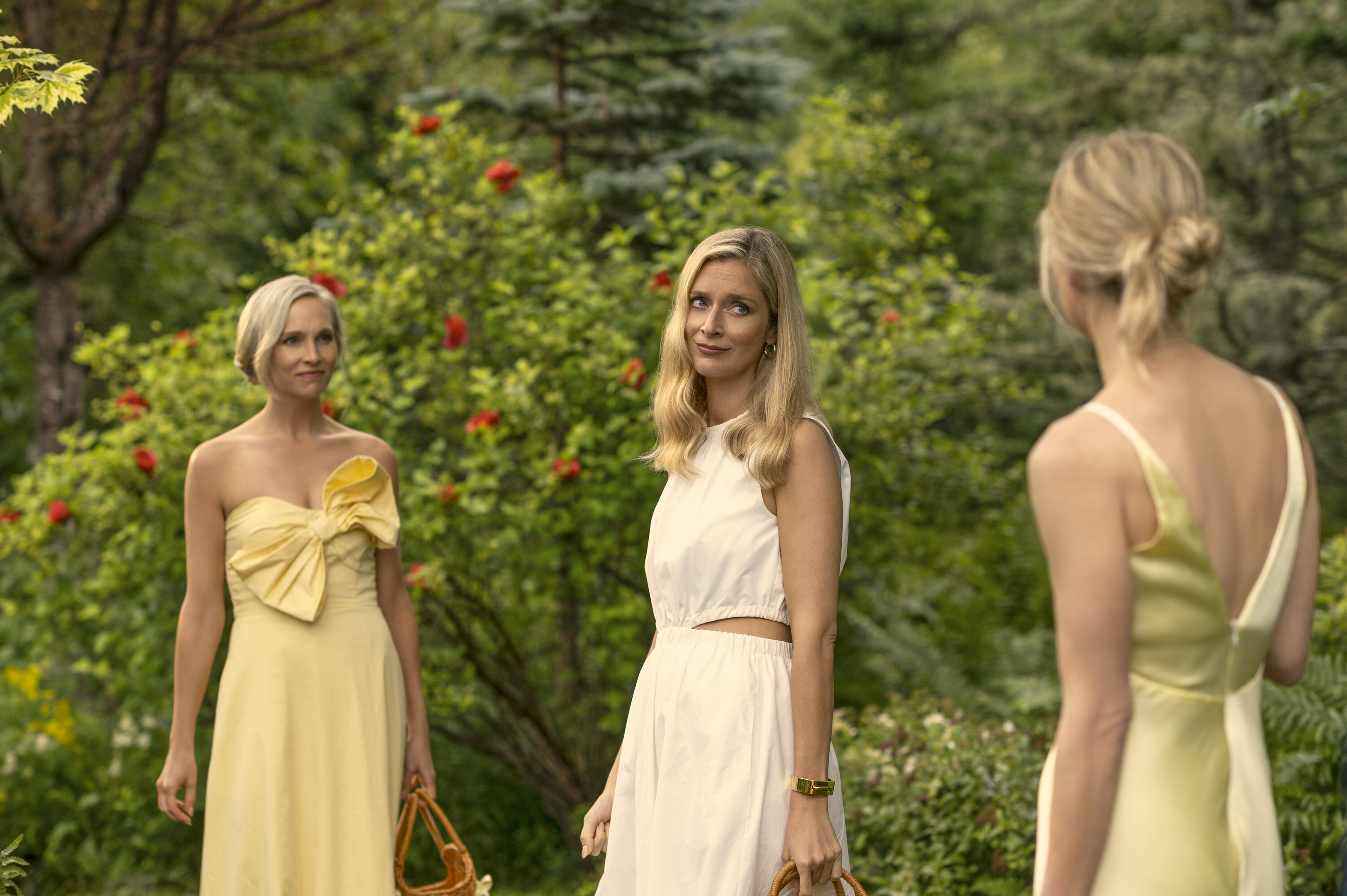 Three white women wearing white and yellow dresses stand in front of greenery, in 'We Were Liars.'
