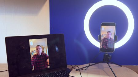 Shot of the AiXPi 10&rdquo; Ring Light holding an iPhone 17. The feed from the iPhone&rsquo;s camera is being displayed on a MacBook. A hand holds the LED&rsquo;s control unit.