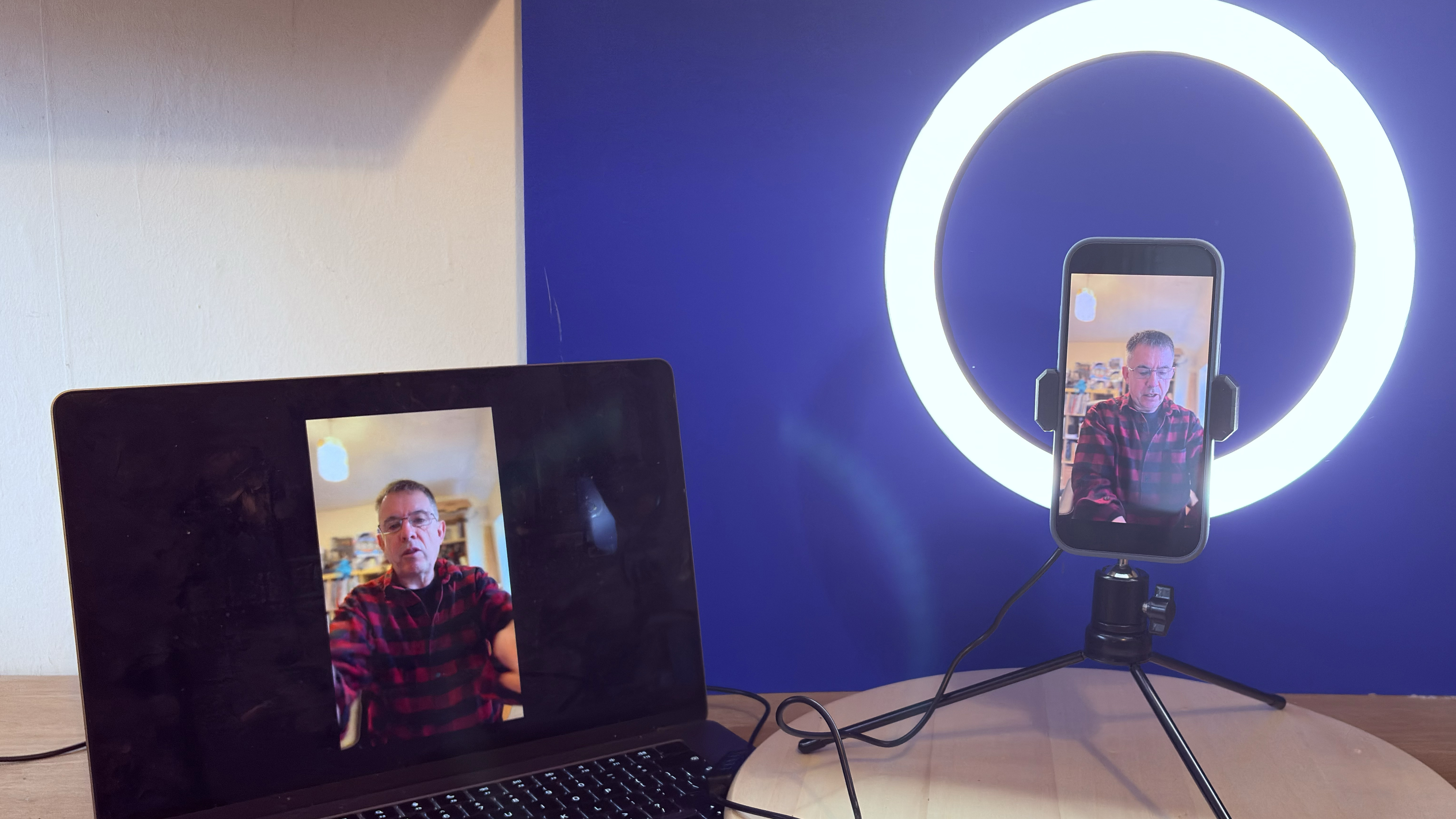 Shot of the AiXPi 10&rdquo; Ring Light holding an iPhone 17. The feed from the iPhone&rsquo;s camera is being displayed on a MacBook. A hand holds the LED&rsquo;s control unit.