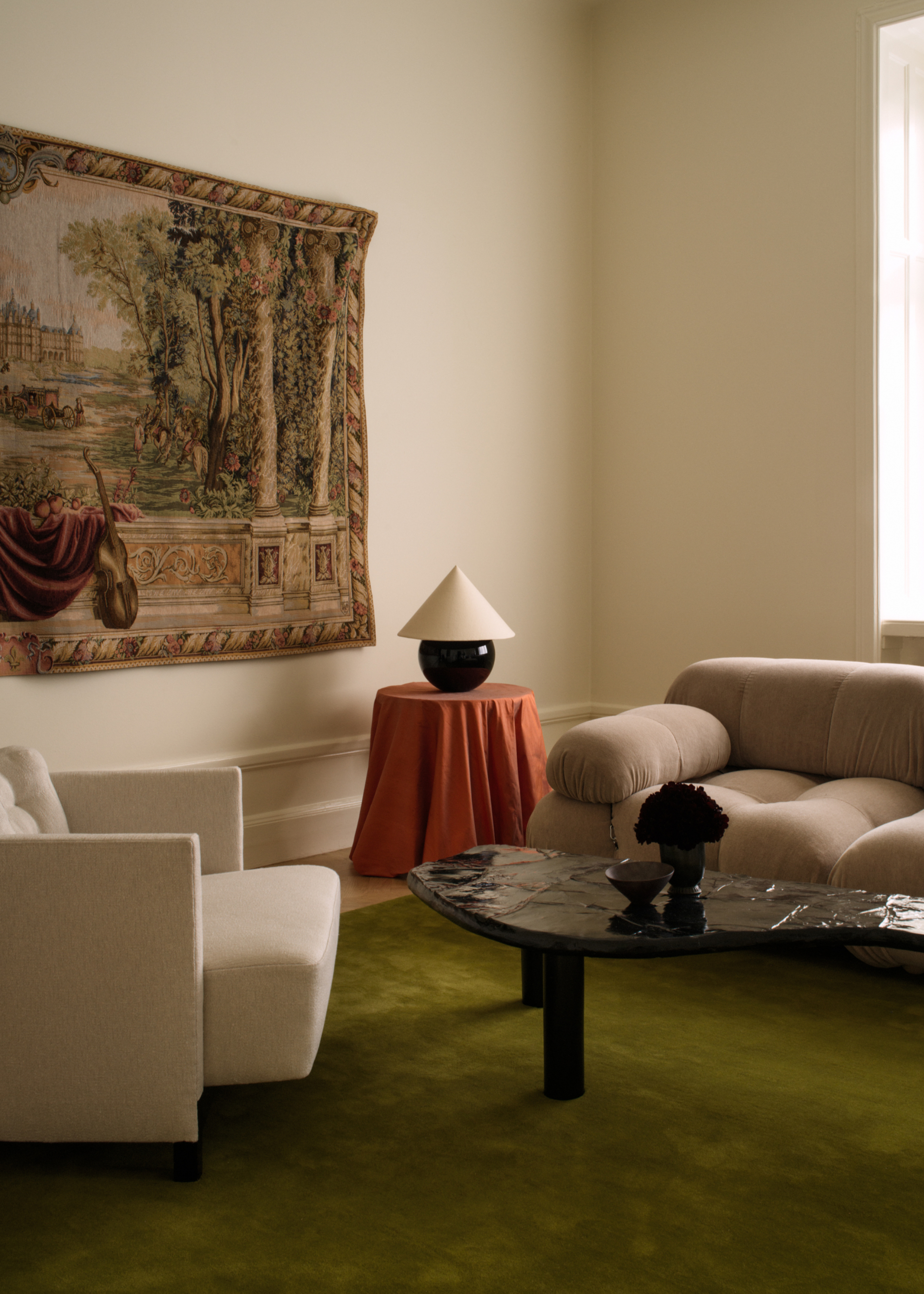 A living room with off-white walls, a printed tapestry, a white armchair, a cream couch, a side table with an orange tablecloth, and a black round and white cone lamp by a black textured coffee table with a vase of flowers and a ceramic bowl on a deep olive green area rug