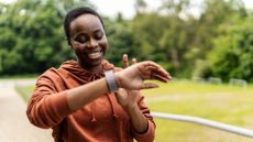 Woman looking at fitness tracker, smiling, standing in a park about to start walking as a workout