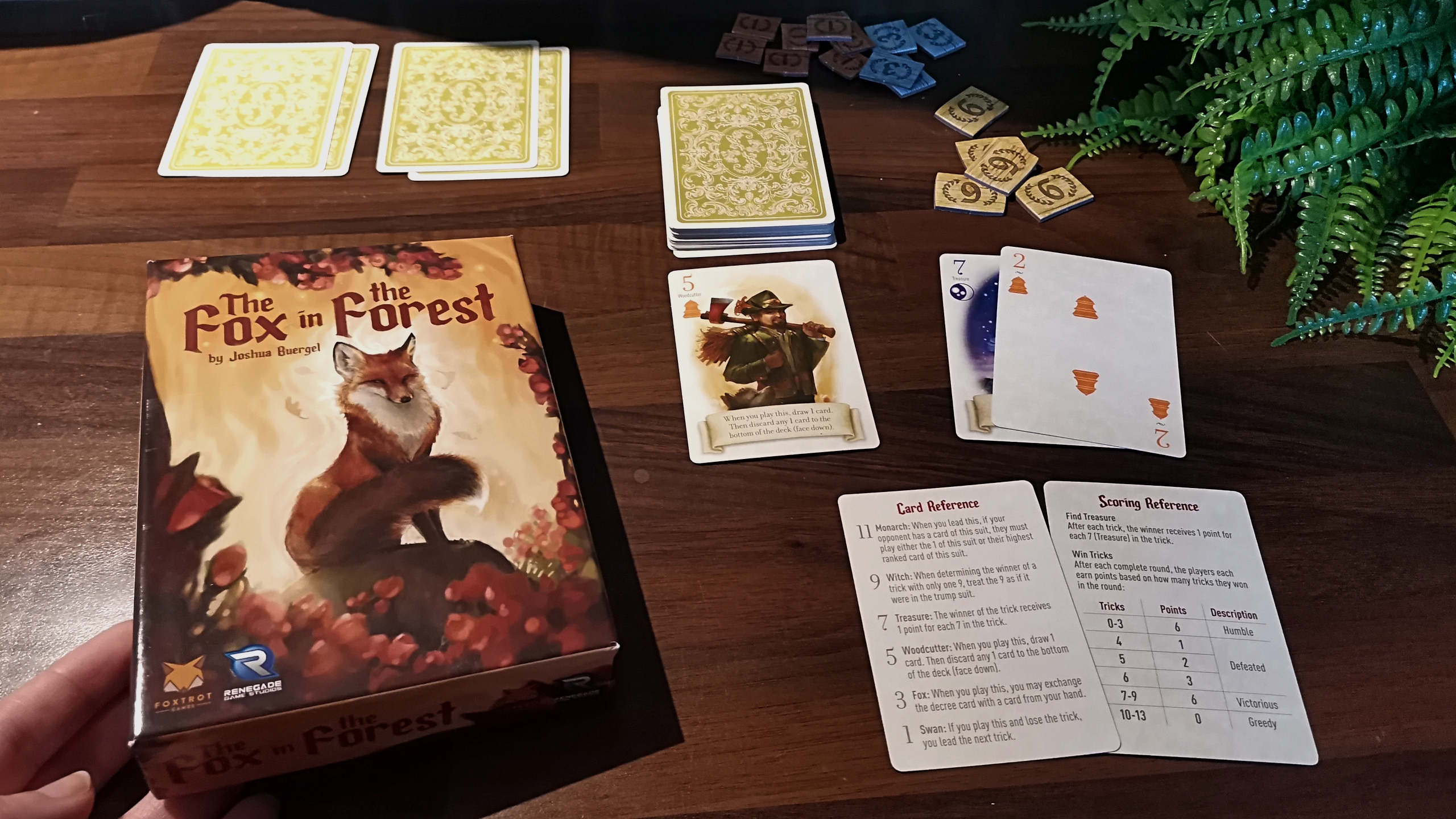 Fox in the Forest box and cards on a wooden table, with a hand reaching toward them from the bottom left corner of the frame