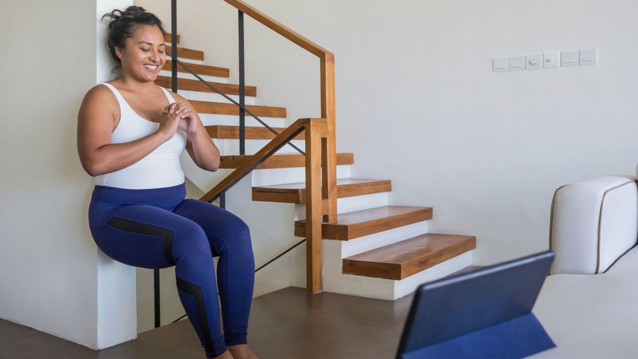 woman doing a wall sit in a home environment in front of a laptop facing the camera, wearing blue leggings and white vest. 