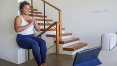 woman doing a wall sit in a home environment in front of a laptop facing the camera, wearing blue leggings and white vest. 