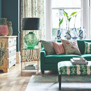 A living room with a green sofa and a botanical-print ottoman for a coffee table and side table with a green glass table lamp