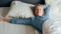 A woman with blond straight hair in a pony tail and wearing blue pajamas stretches on a bed with white sheets and pillows, smiling with her eyes closed