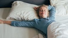 A woman with blond straight hair in a pony tail and wearing blue pajamas stretches on a bed with white sheets and pillows, smiling with her eyes closed