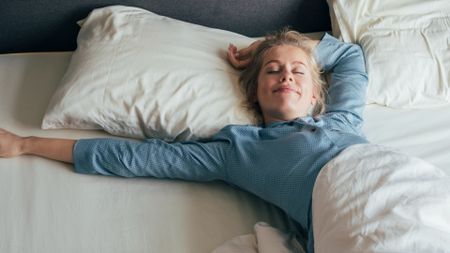 A woman with blond straight hair in a pony tail and wearing blue pajamas stretches on a bed with white sheets and pillows, smiling with her eyes closed