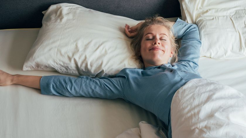A woman with blond straight hair in a pony tail and wearing blue pajamas stretches on a bed with white sheets and pillows, smiling with her eyes closed