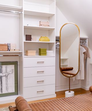 A white closet room with shelves, a set of drawers, a full-length curved mirror in front of a hidden wall storage door, and a light leather brown bench with curved cushion arms