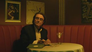 Ian McShane sits writing at a table with a martini nearby in John Wick.