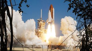 a white and black winged spacecraft attached to white boosters and an orange-brown tank lifts off from a launch pad framed by trees in silhouette in the foreground