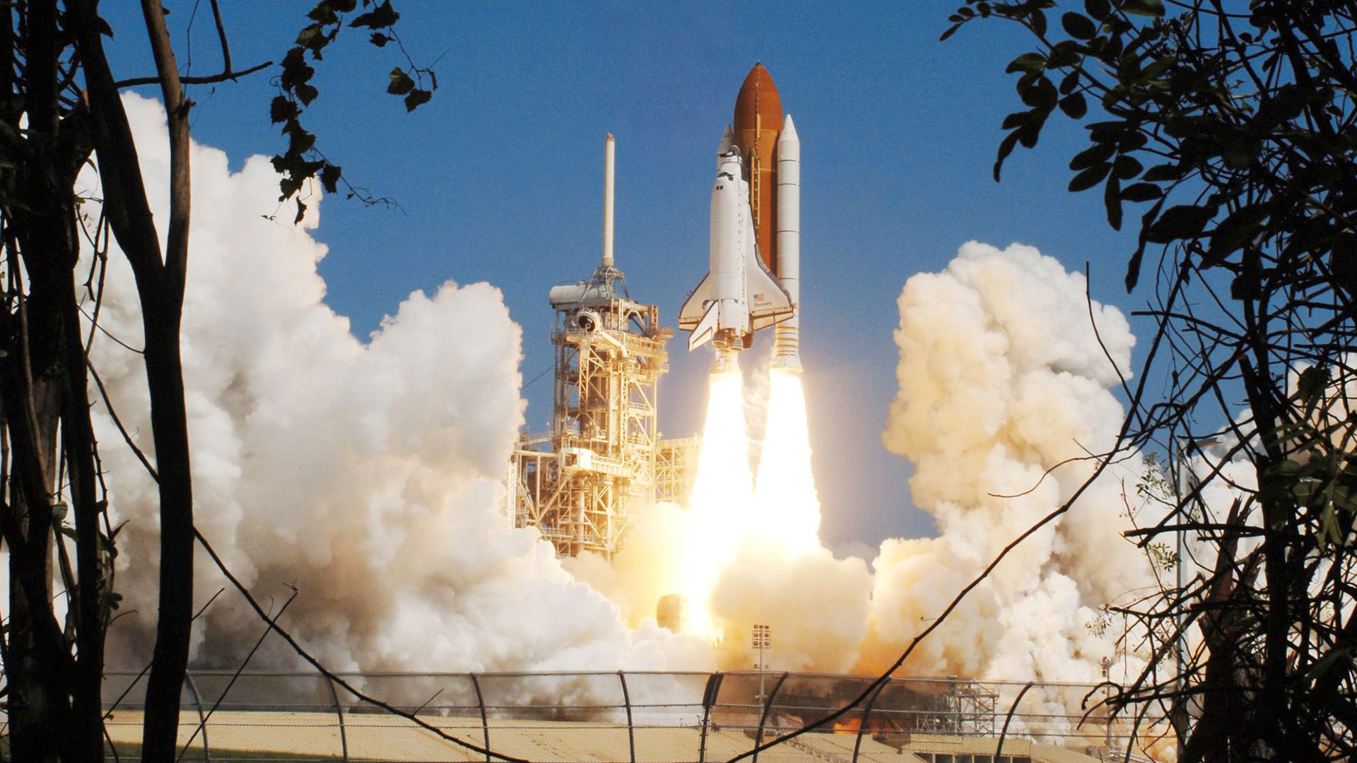 a white and black winged spacecraft attached to white boosters and an orange-brown tank lifts off from a launch pad framed by trees in silhouette in the foreground