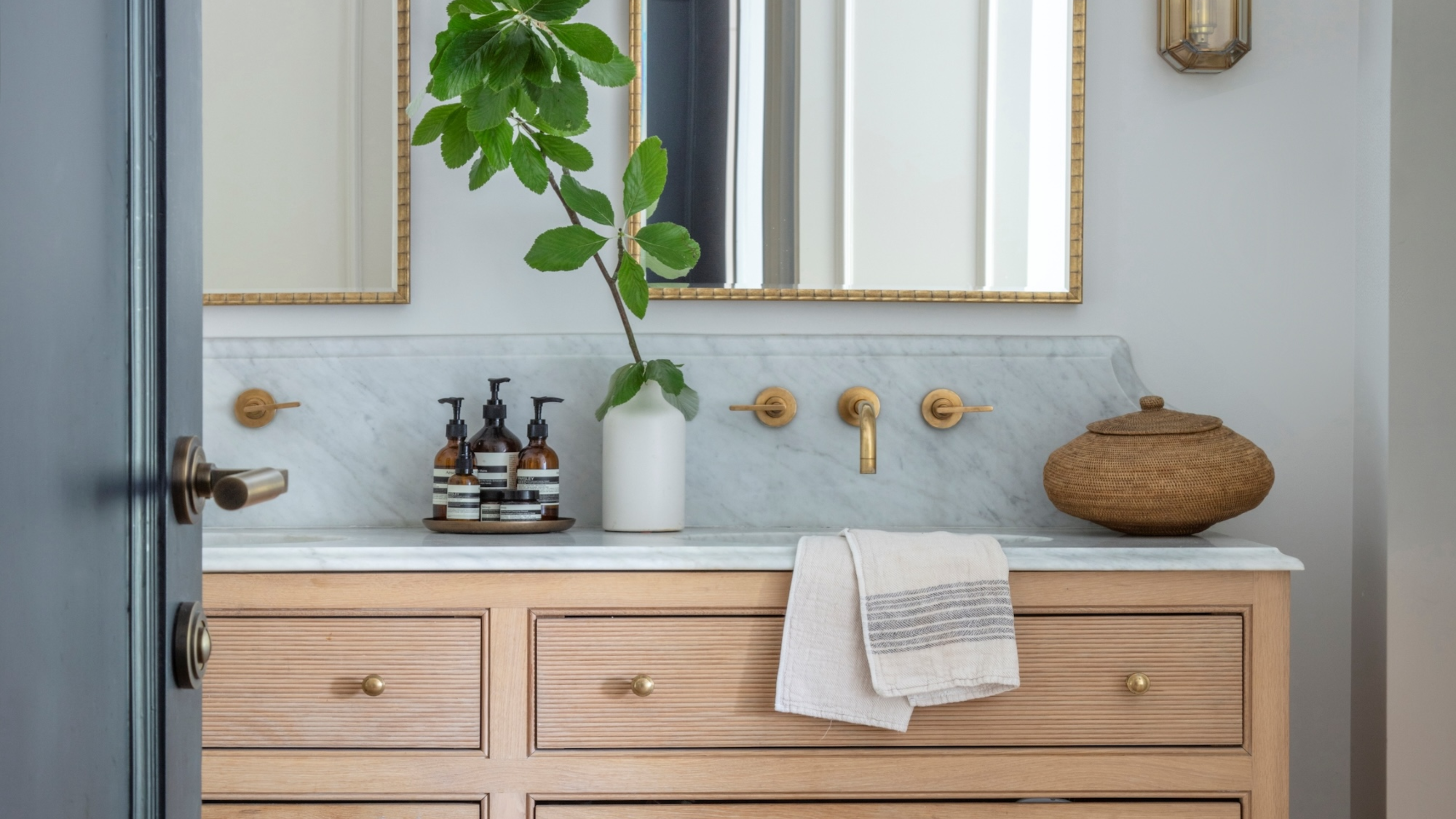 A guest bathroom with a vanity On top of the vanity is a tray of body wash/ soap as well as a plant in a vase and a small rattan basket. Above the vanity are two large bobbin mirrors. 