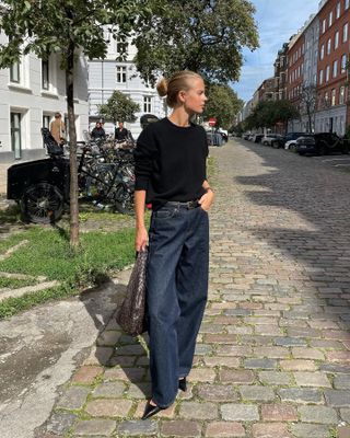 Swedish art director Clara Dyrhauge wears a stylish outfit with a black sweater, skinny belt, woven bag, dark wash jeans, and pointed-toe heels.