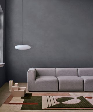Living space with mottled graphite painted wall, large gray sofa, geometric rug in muted colorways, wooden decorative object on floor, low hanging pendant with opal glass rounded shade
