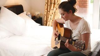 Girl sat on bed with an acoustic guitar looking down at a book with notes in. 