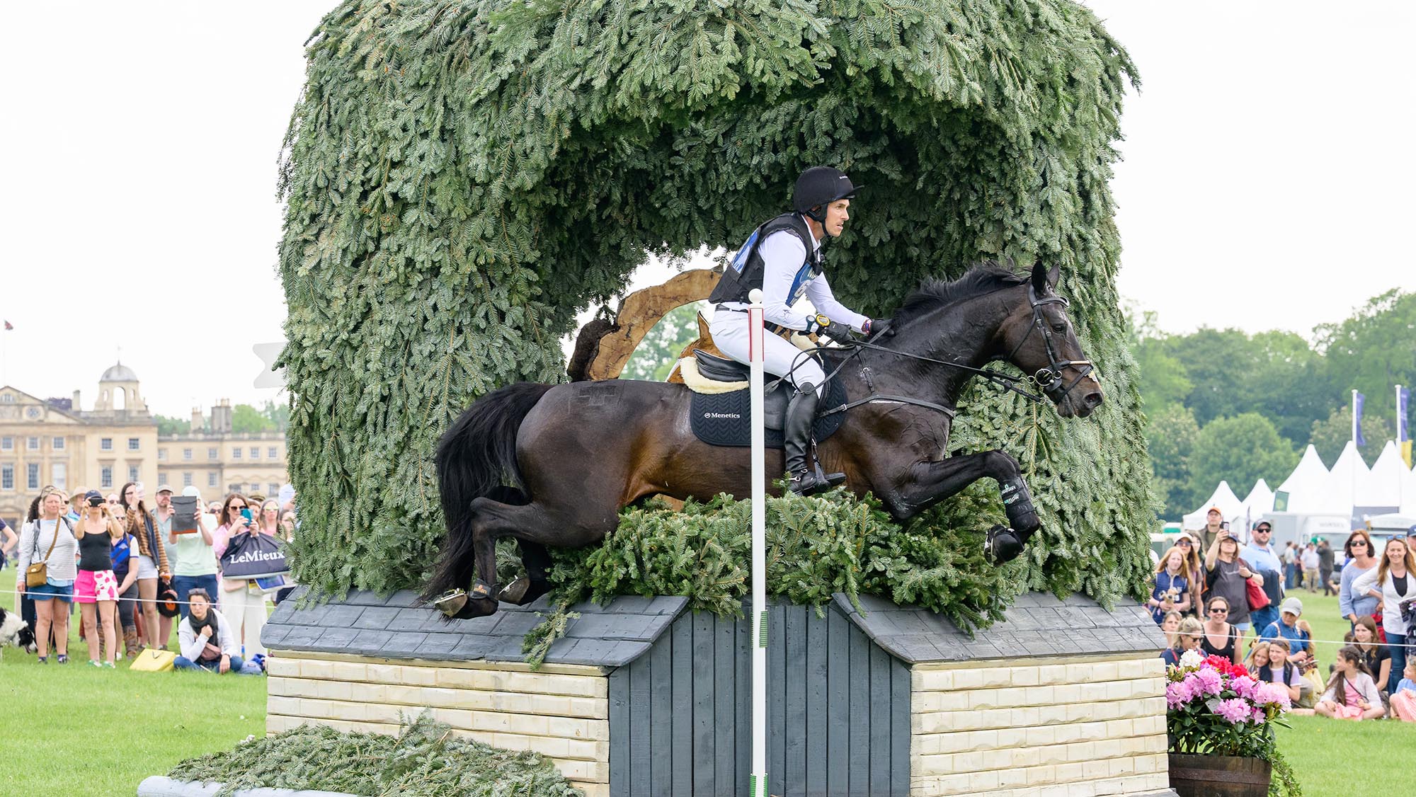 Felix Vogg, a Swiss rider, rides Cartania, a dark brown horse, over a corner jump with brush over the top of it, in front of Badminton House