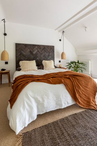 A loft bedroom with DIY headboard, two rattan ceiling pendant sides on either side of bed, rust colored throw on bed, cream carpet and grey-brown runner rug
