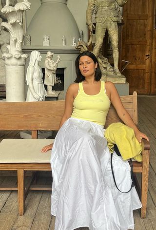 A woman wearing a yellow tank top with a white maxi skirt, flat sandals, a yellow cardigan, and a black bag.