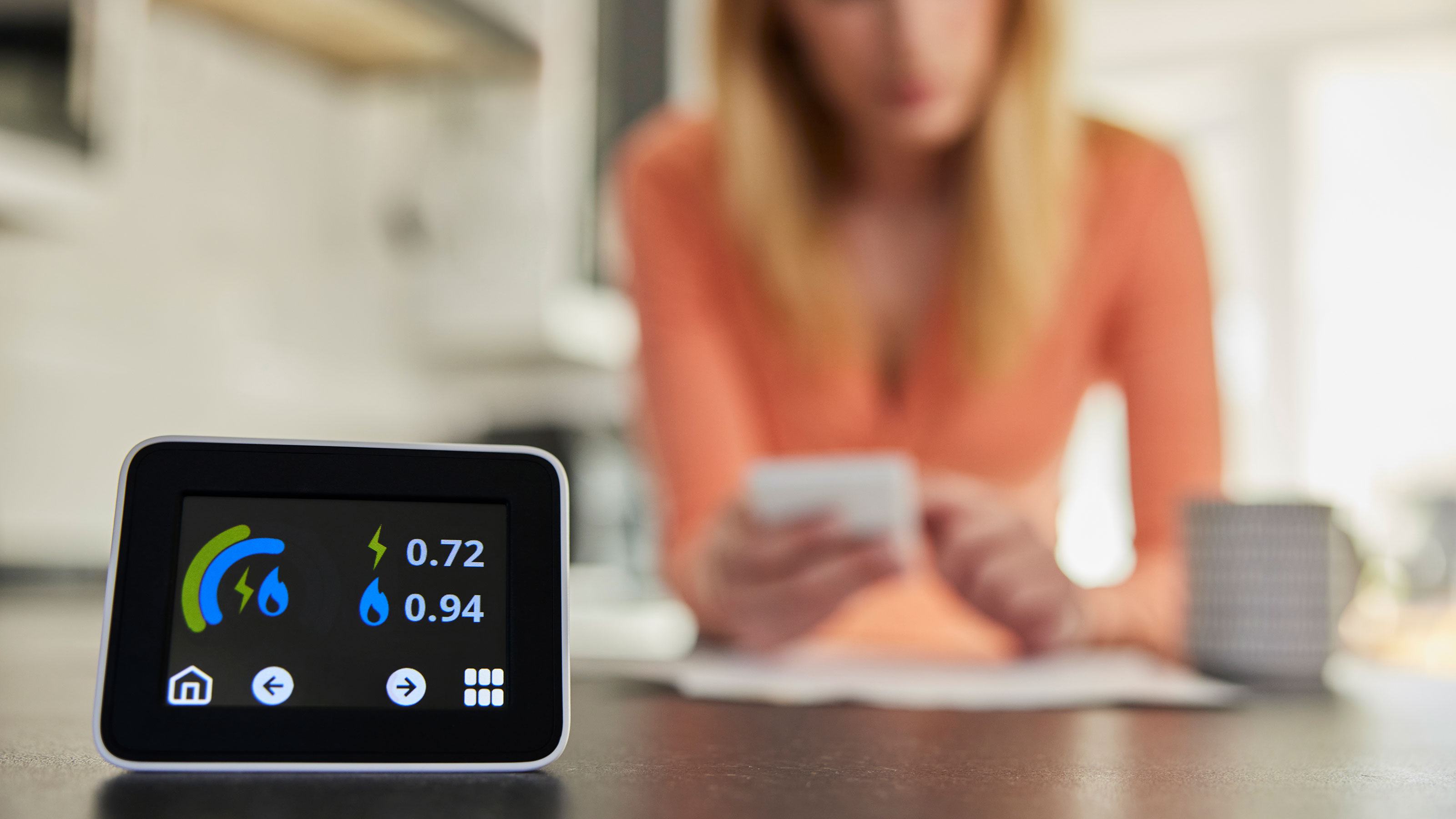 Close Up Of Smart Energy Meter In Kitchen Measuring Electricity And Gas Use With Woman Looking At Bills With Calculator