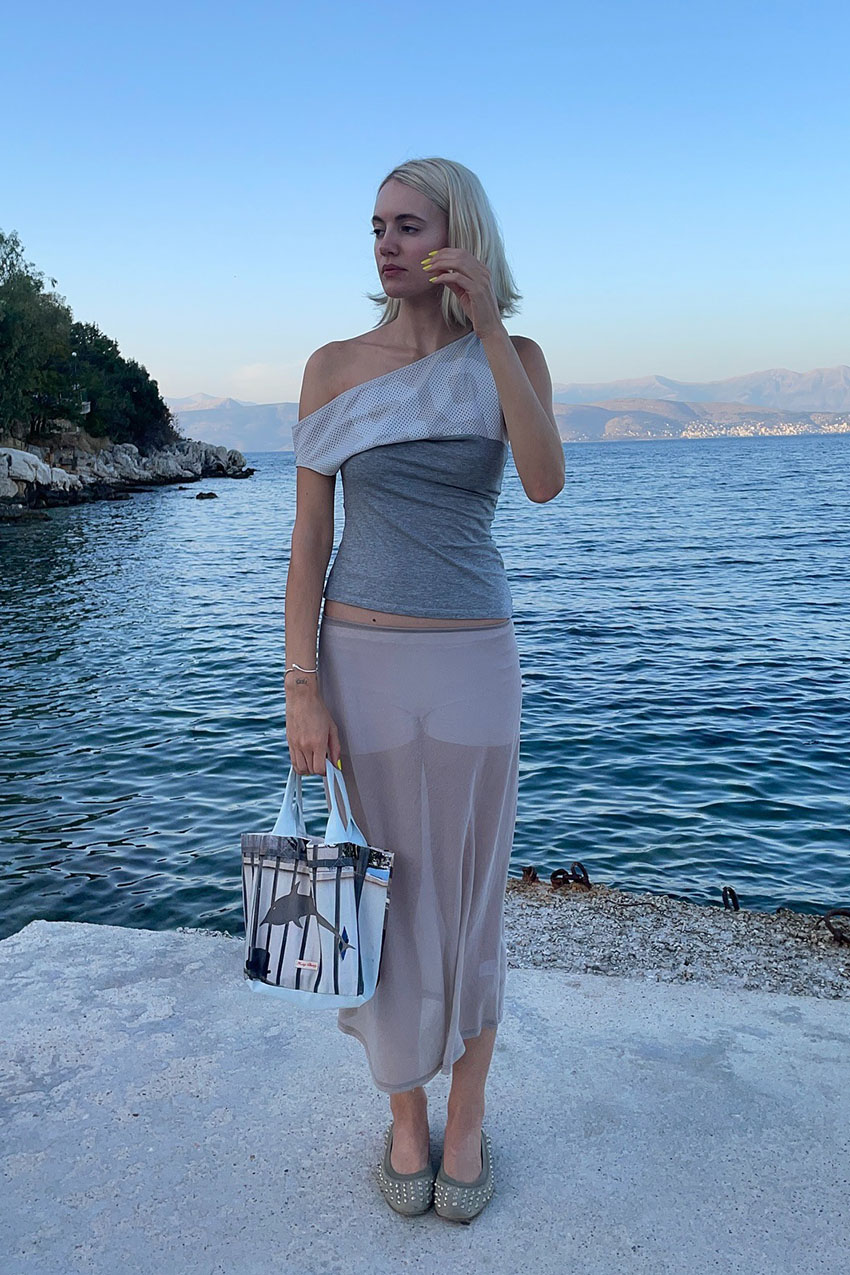 a woman wearing a one shoulder top, sheer tan skirt, studded flats, and a graphic tote bag