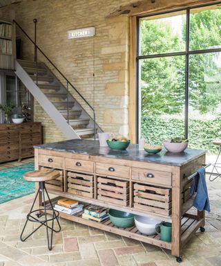 A portable rustic-industrial kitchen island on wheels with blue and gray bowls above it and beneath it and a staircase and window behind it