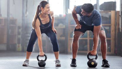 A man and a woman are working out in a gym next to each other. He wears shorts and a t-shirt with sneakers, she wears a vest, mid-calf leggings and sneakers. Her long hair is tied back in a pony tail. They are both squatting down, with their knees slightly bent, and one arm hanging in front of them grasping the handle of a kettlebell weight. Their other hand rests on their hip. They look towards each other and the woman is smiling.