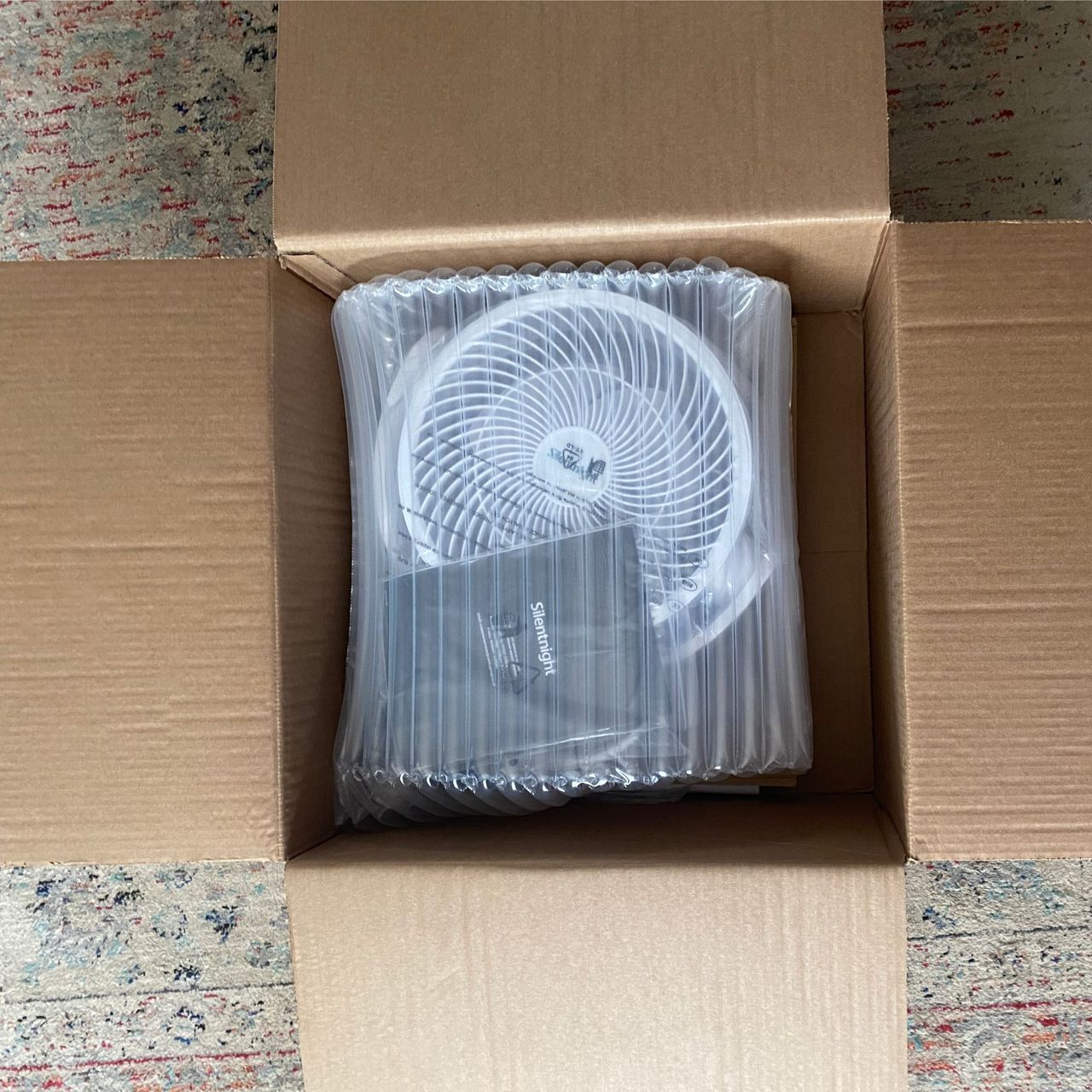 The inflated internal packaging of the Silentnight Airmax Pro360 Pedestal Fan, in an open cardboard box, on the floor on a patterned rug