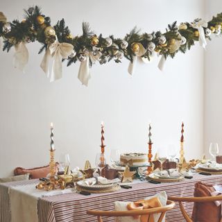 A dining table set for a Christmas dinner with a garland hanging above decorated with metallic baubles and white satin bows