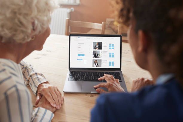 Mature female nurse teaching senior woman booking appointment on laptop at home