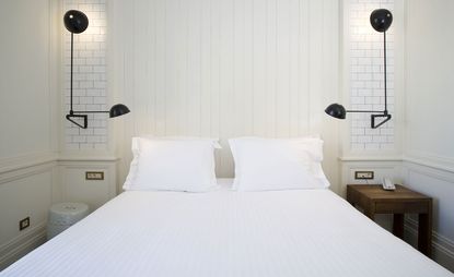 A hotel bedroom with a bed, a wooden side table, black wall lamps, a round grey stool and white wood decorated walls.