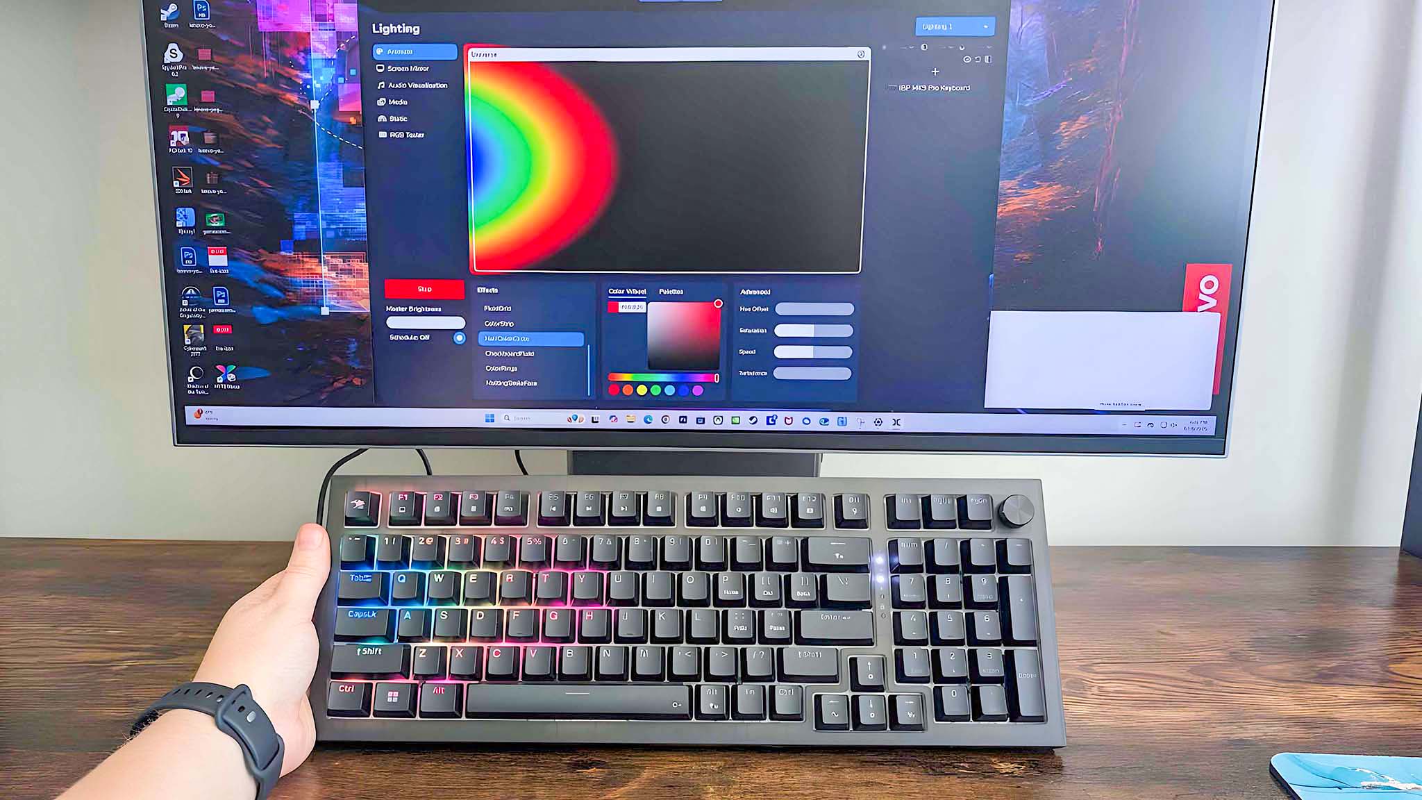 A hand holding the iBuyPower MK9 Pro RGB Mechanical Keyboard up and showing that the keyboard's rainbow RGB effect matches what's shown on the computer's screen in the back.