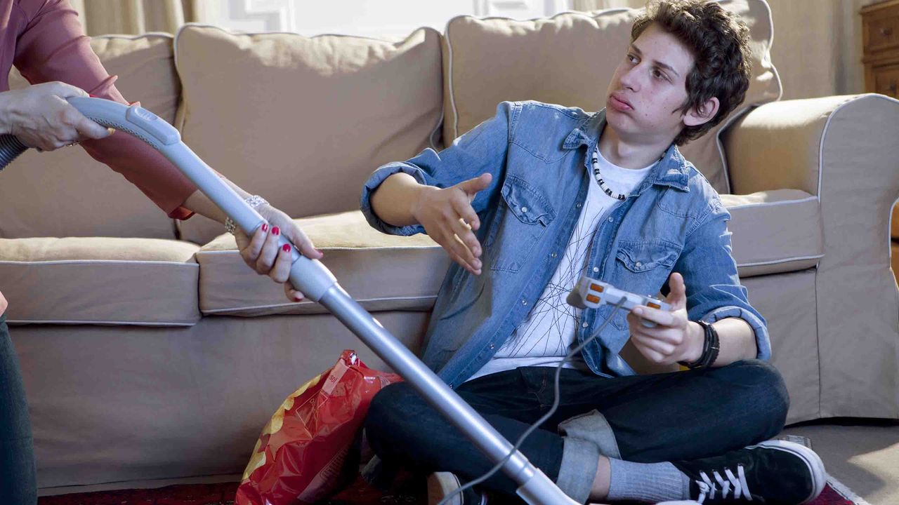 Photo of a young adult male sitting on the floor looking at a phone while someone is vacuuming around him. 