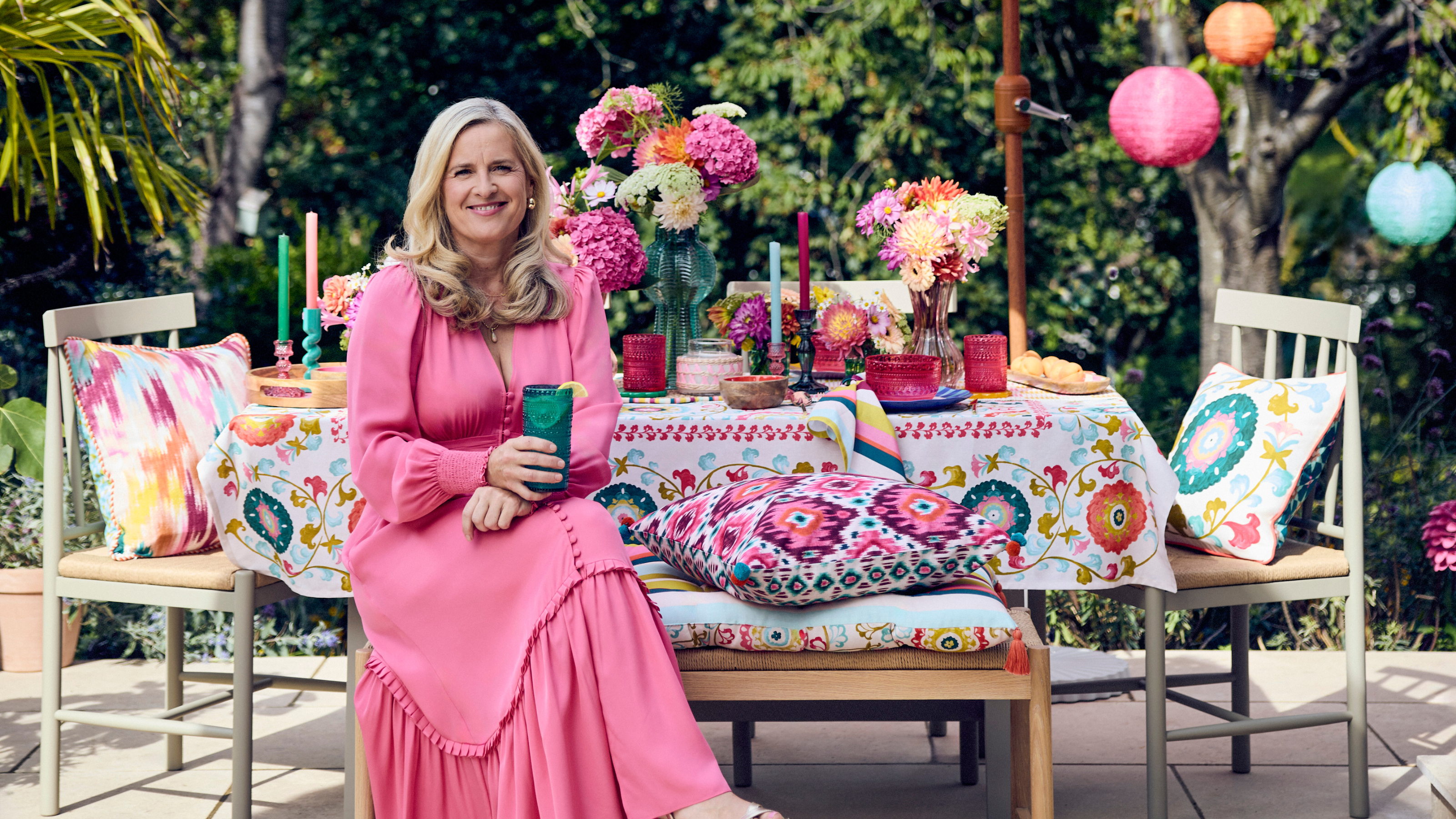 Sophie Robinson in pink dress sat at bench decorated with colourful candles, tablecloth and flowers in a patio garden.